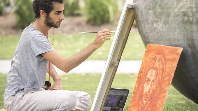 Student with headphones sits and paints outside on a canvas.