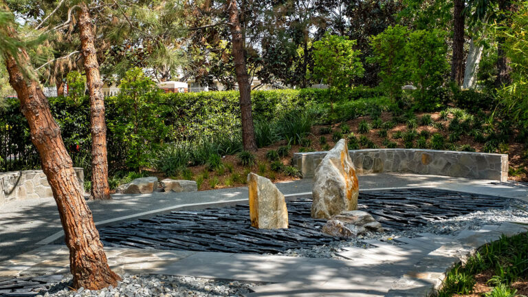 The serene and quiet Nisei rock garden, with two large stones in the middle surrounded by flatter rocks in layers - almost looking like two rocks floating in the ocean. Pine trees and greenery surrounds the rocks, providing peaceful shade.
