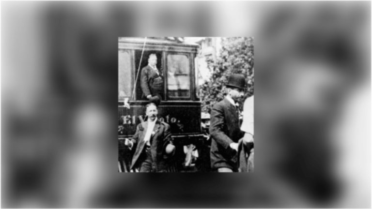 US President Taft stands in the back of a train car, looking out the open window toward a group of onlookers in the foreground