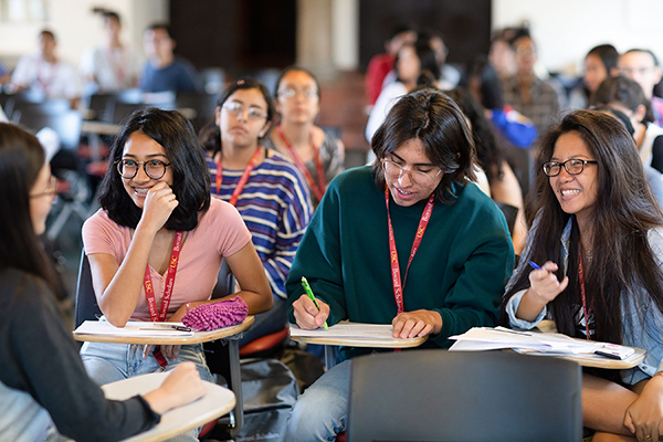 Smiling students wearing lanyards sit at desks and socialize.