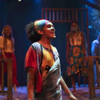 An actor singing, wearing a bandana and overalls, in a USC School of Dramatic Arts stage performance of 