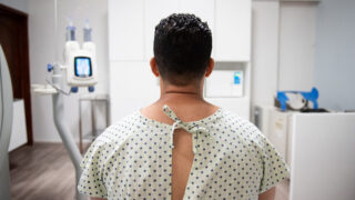 Patient’s shoulders and head are seen from behind. He is wearing a hospital gown.